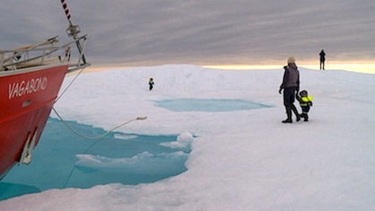 Photo : surtout de la glace et de l'eau très bleu, une mère, deux bébés et un homme au loin se promènent, vêtus en habits d'hiver moderne. À gauche, on voit la pointe du bateau rouge nommé Vagabond.